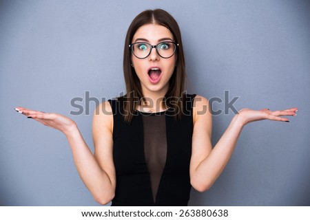 Young beautiful woman with facial expression of surprise standing over gray background. Wearing in trendy black dress and glasses. Looking at the camera