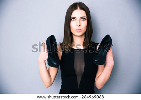 Portrait of a surprised cute woman holding shoes. Wearing in black dress. Looking at camera.