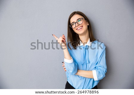 Cheerful businesswoman pointing away over gray background. Wearing in blue shirt and glasses. Looking away