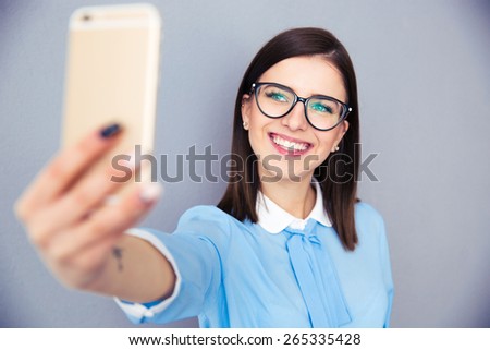 Smiling businesswoman making selfie photo on smartphone. Wearing in blue shirt and glasses. Standing over gray background