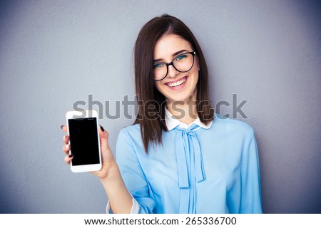 Smiling businesswoman showing blank smartphone screen over gray background. Wearing in blue shirt and glasses. Looking at camera.