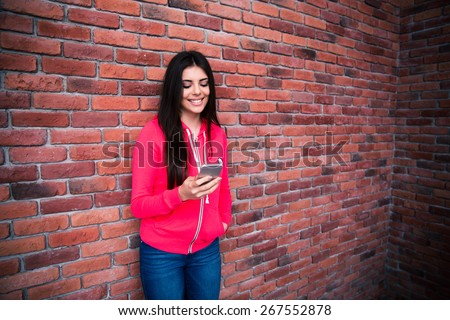 Young smiling woman using smartphone over brick wall.