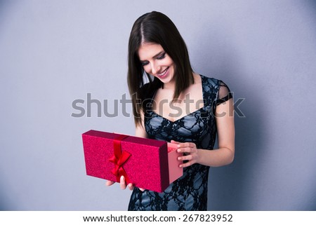 Happy young woman opening gift box and looking on present over gray background