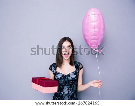 Cheerful woman holding gift box and balloon over gray background. Wearing in dress. Looking at camera