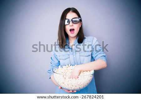 Surprised pretty woman in cinema glasses eating popcorn over gray background. Looking at camera