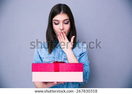 Surprised young woman standing with gift over gray background. Looking on gift. Covering mouth with her hand