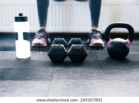 Closeup image of a woman sitting at gym with dumbbells, shaker and weight