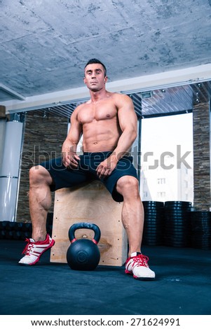 Muscular man sitting on fit box and reading for workout with kettle ball in gym. Looking at camera