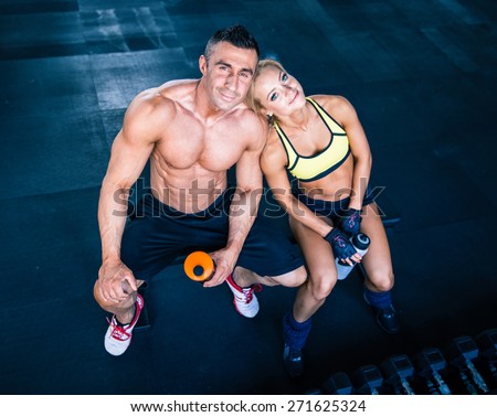 Smiling muscular man and happy sporty woman resting on bench at gym. Looking at camera