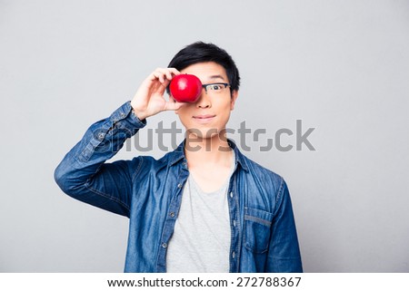 Young asian man covering his eyes with apple over gray background. Looking at camera