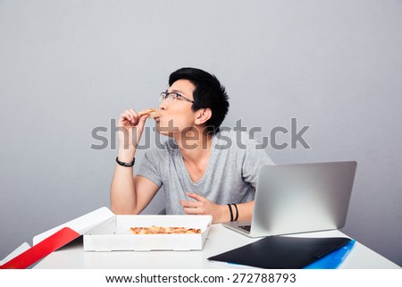 Handsome asian man sitting at the table and eating pizza over gray background