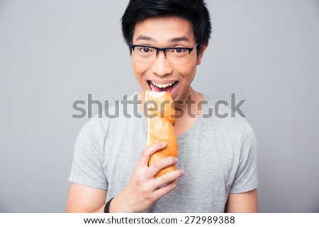 Asian man in glasses biting bread over gray background and looking at camera