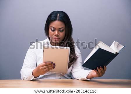 African businesswoman choosing between e-book or paper book over gray background. Looking at camera