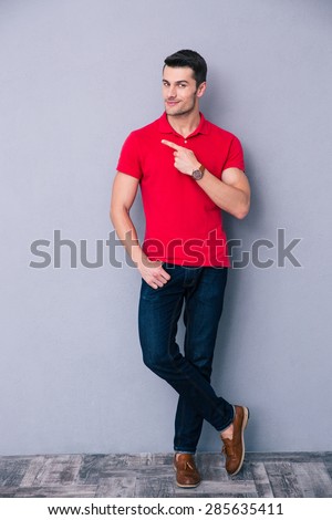 Full length portrait of a casual man pointing away and leaning on the gray wall