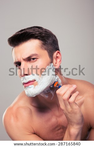 Young handsome man shaving with foam and razor over gray background