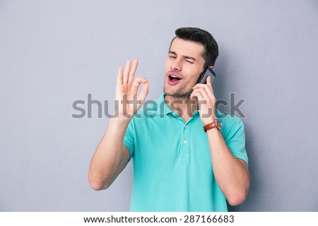 Happy young man talking on the phone and gesturing ok sign over gray background