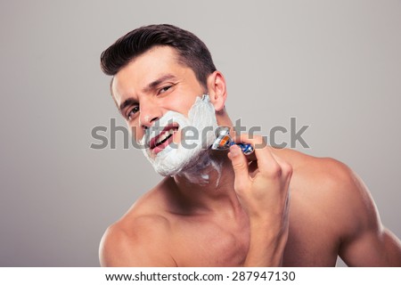 Young man shaving with foam and razor over gray background