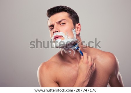 Young man shaving with foam and razor over gray background