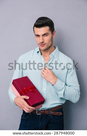 Handsome young man holding gift box over gray background. Looking at camera