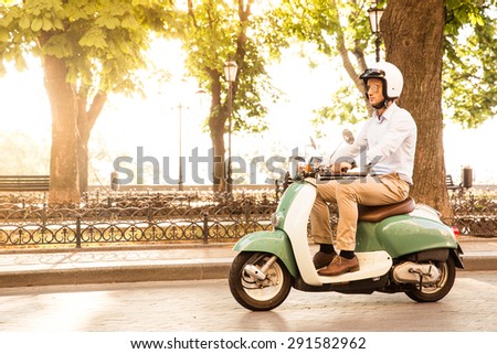 Trendy man driving a scooter in helmet. Sun is shining through trees on background
