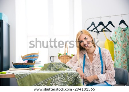 Happy female tailor sitting at her workplace in workshop and looking at camera