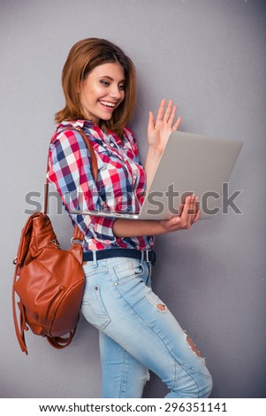 Happy young woman standing with laptop and showing greeting gesture to web camera over gray background