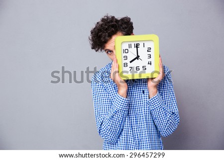 Man covering his face with big clock and looking at camera over gray background