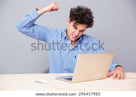 Angry man sitting at the table with laptop over gray background