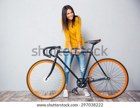 Full length portrait of a smiling woman standing near bicycle on gray background. Looking at camera