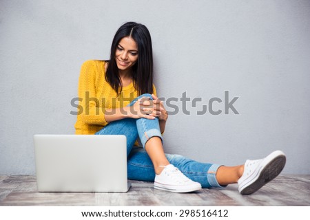Smiling young woman sitting on the floor with laptop on gray background