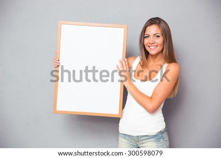 Happy casual woman holding blank board over gray background. Looking at camera