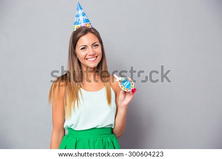 Happy young girl in birthday hat and whistle standing over gray background and looking at camera