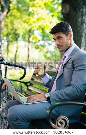 Portrait of a handsome businessman sitting on the bench with laptop and drinking coffee outdoors