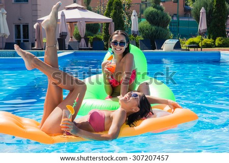 Two funny girls lying on air mattress in swimming pool with cocktails