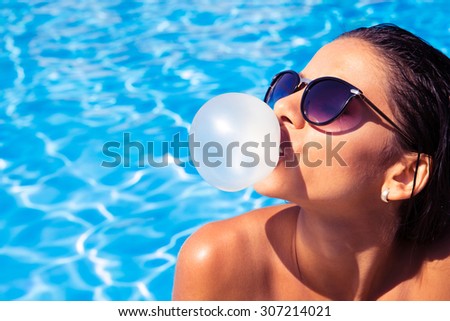 Charming woman in sunglasses blowing bubble with gum in swim pool outdoors
