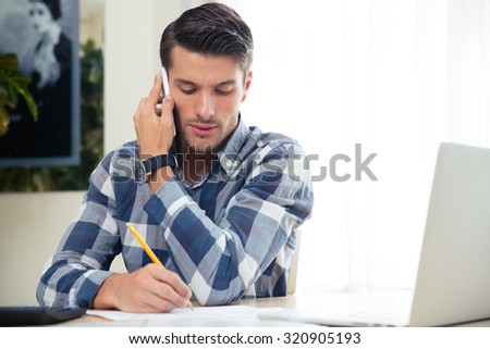 Portrait of a man making notes on the bills while talking on the phone at home