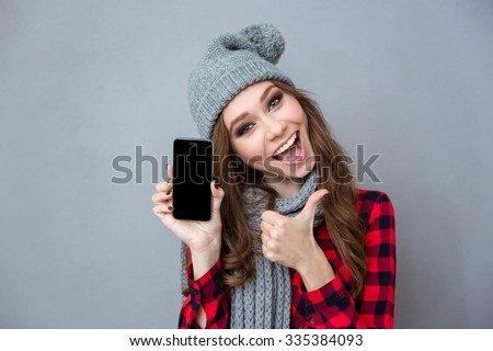 Portrait of a cheerful woman showing blank smartphone screen and thumb up over gray background