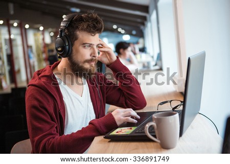 Handsome serious boy with beard in brown hoodie using laptop and listening to music