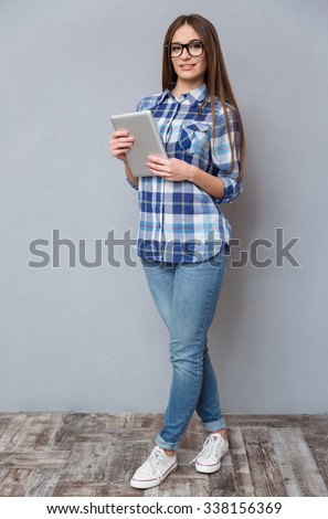Full length portrait of beautiful happy smiling smart girl in checkered shirt and glasses holding tablet