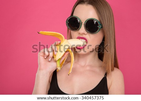 Closeup of young seductive attractive sensual model in black sunglasses eating banana on pink background