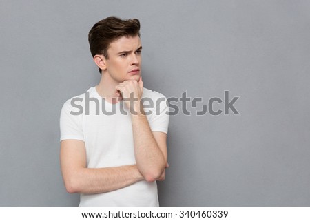 Portrait of pensive serious concentrated smug young man in white t-shirt posing on gray background