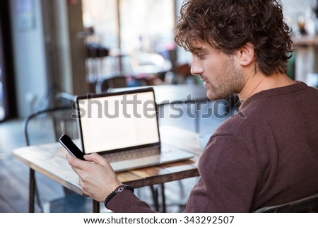 Back view of handsome curly concentrated attractive man using cellphone while working with laptop