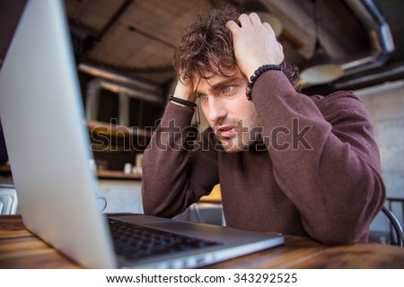 Stressful upset desperate handsome curly man in brown sweetshirt working using laptop and having headache