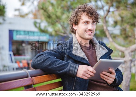 Curly positive handsome young guy on black jacket using tablet on the wooden bench in park and looking away