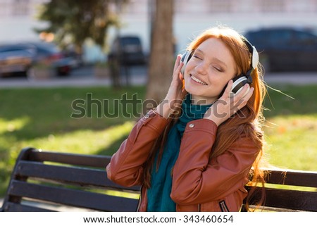 Content cheerful young woman in leather jacket and scarf sitting on bench in park and listening to music with eyes closed