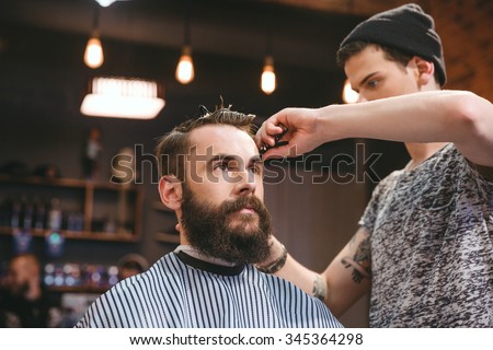 Handsome skillful barber cutting hair of young attractive man with beard at barbershop