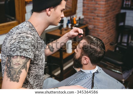 Young hairstylist with tattoo combing hair of male client with a comb in hair salon