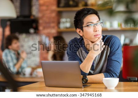Thoughtful handsome young asian man in glasses sitting with laptop in cafe and looking away