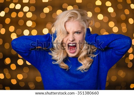 Portrait of a blonde woman covering her ears and screaming over holidays lights background