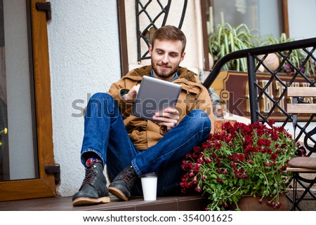 Smiling bearded young man in coat and jeans sitting on porch near entrance using tablet